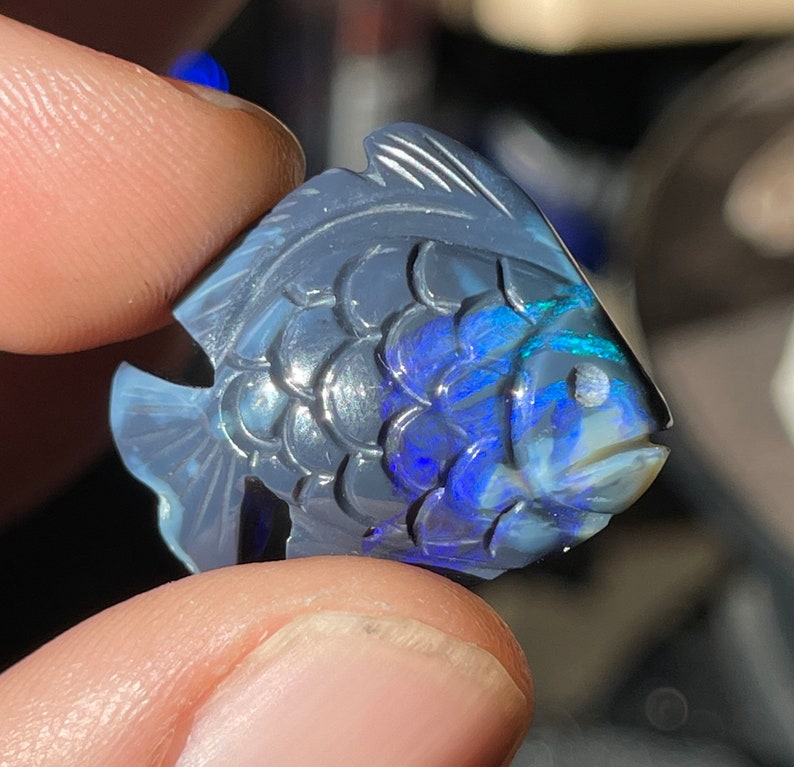 Hand-carved opal fish with detailed scales — translucent blue body with vivid blue and green play-of-color inside, held in the cutter's fingers.