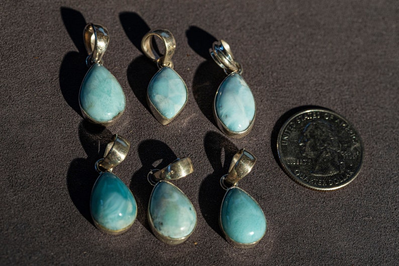 Six finished Larimar pendants in sterling silver bezel settings, photographed next to a US quarter for scale — pale blue stones with subtle pattern.