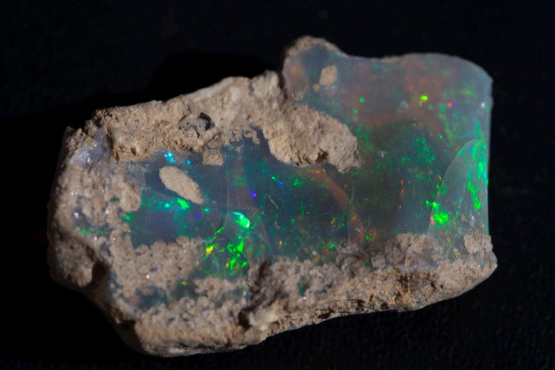 Rough Ethiopian Welo opal in cream-colored host matrix — vivid green and blue play-of-color glowing through the gem pocket.
