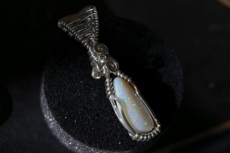 Wire-wrapped opal pendant in fine sterling silver — the wire forms a delicate spiral cradle around a long opal cabochon, photographed on black velvet.
